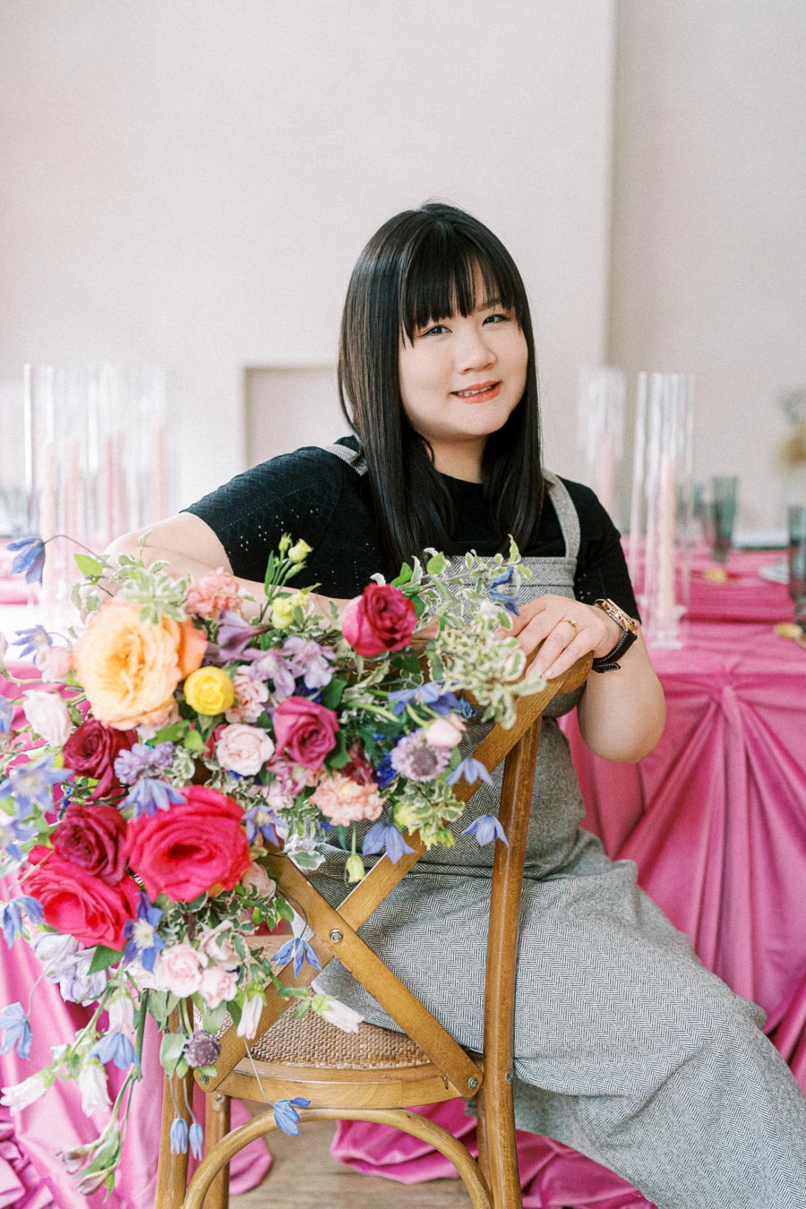 A woman with long dark hair smiling while seated on a wooden chair, holding a vibrant bouquet of colorful flowers, with a pink draped table in the background.