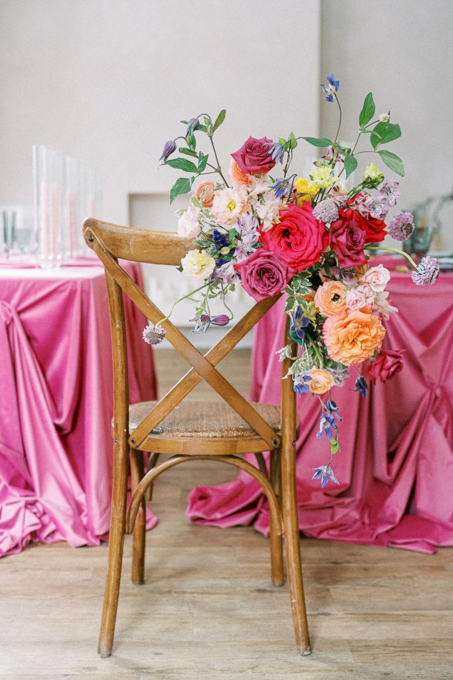 Wooden chair adorned with vibrant floral arrangement featuring roses and mixed blossoms, set against a pink draped table at a wedding or event venue.