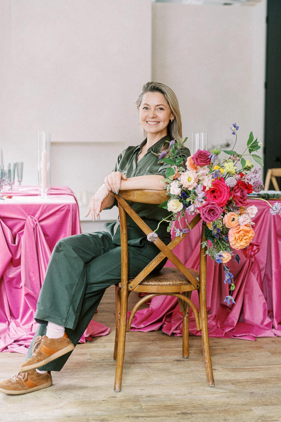 A smiling person sitting on a wooden chair adorned with vibrant, colorful flowers, with a table draped in bright pink fabric in the background, creating a cheerful and elegant setting.