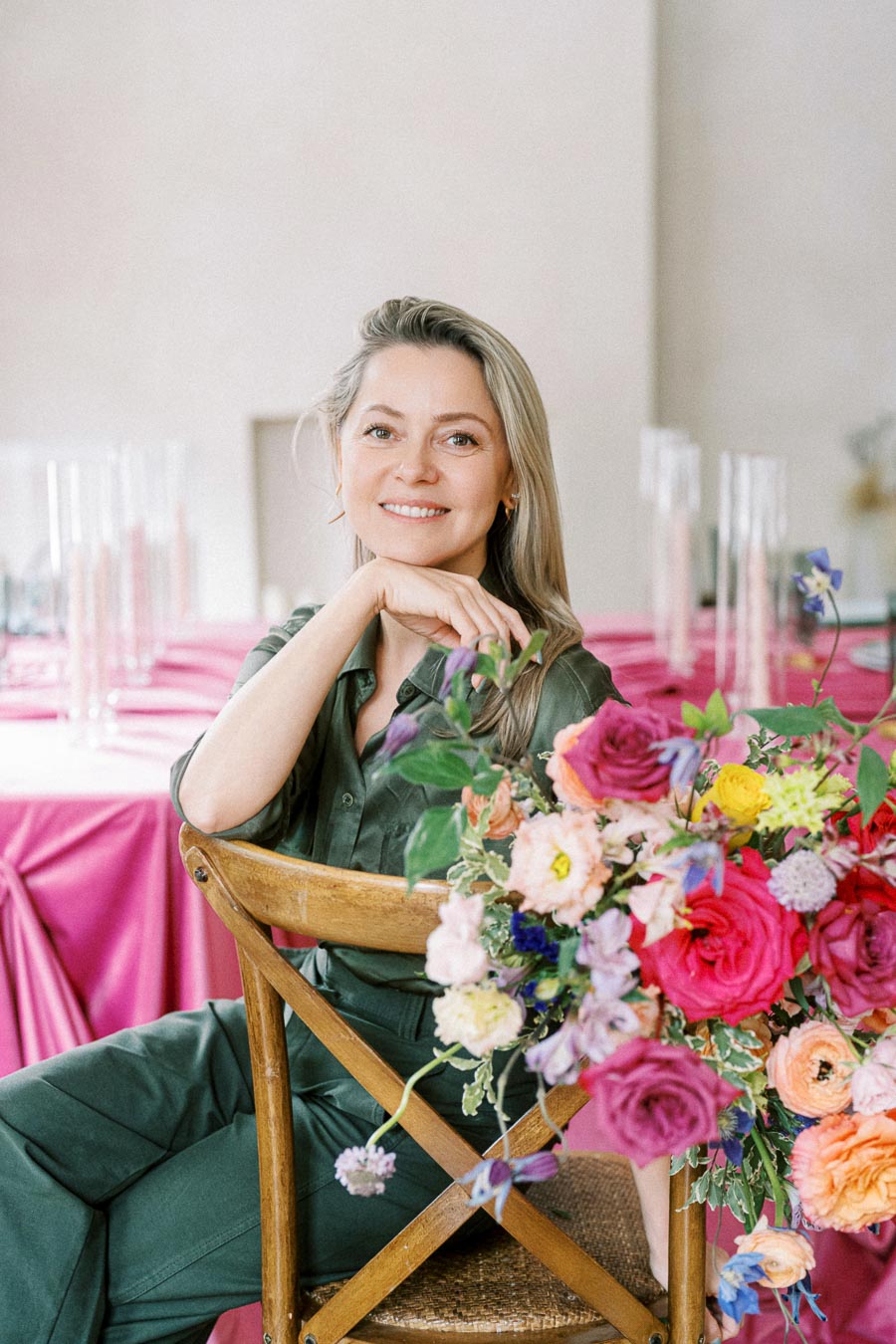 A smiling woman in a green outfit sits on a wooden chair, surrounded by vibrant, colorful flowers. She poses in a room with tables covered in pink tablecloths, creating a cheerful and elegant atmosphere.