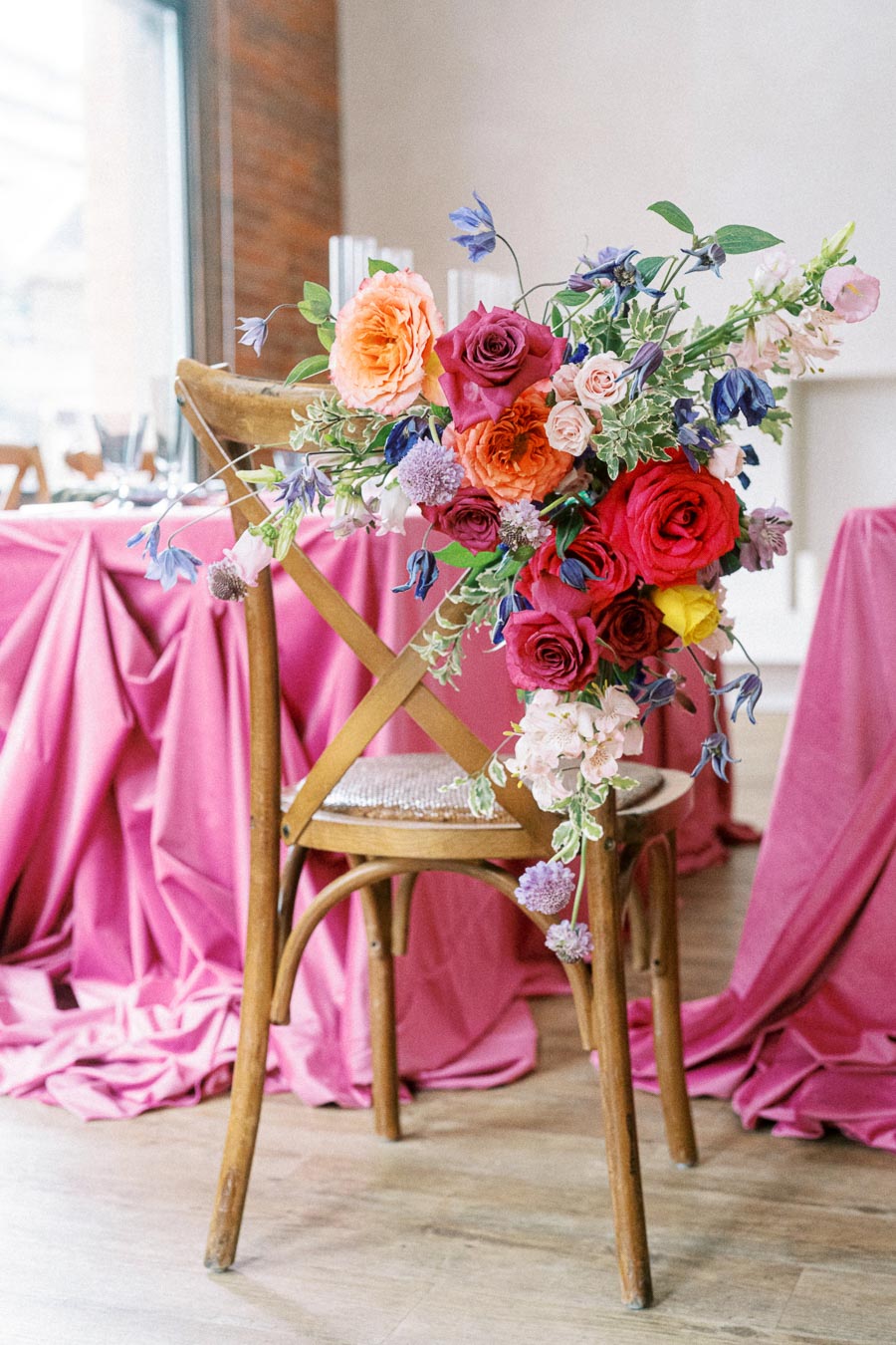 Elegant wedding decor featuring a wooden chair adorned with a vibrant floral arrangement of red, orange, and purple roses, set against tables draped in pink cloths. Perfect for romantic event ambiance.