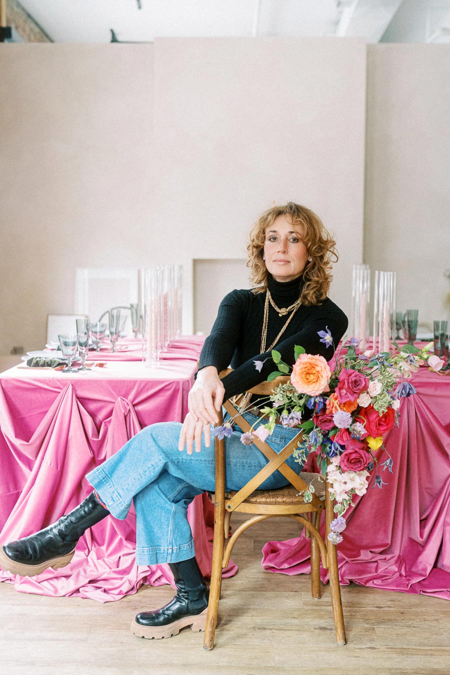 A woman sitting on a wooden chair in front of a table with a pink tablecloth, decorated with elegant glassware and colorful floral arrangements. She is wearing a black turtleneck, jeans, and black boots, with a confident expression. The setting suggests a sophisticated event or gathering.