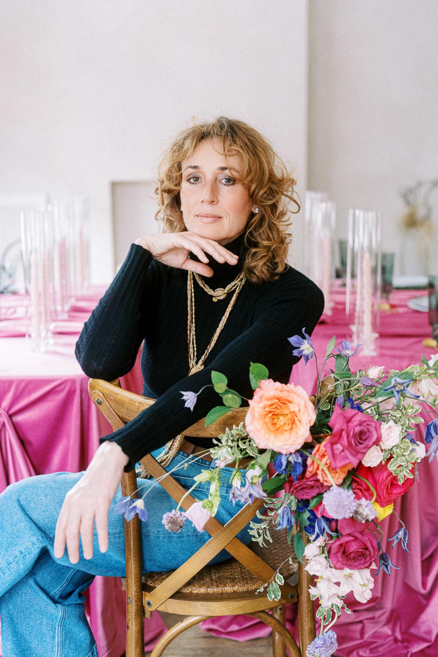 Woman seated on a wooden chair with a colorful floral arrangement, wearing a black turtleneck and jeans, against a backdrop of pink tablecloths and glass vases.