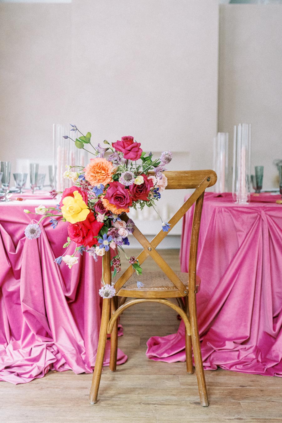 Elegant wedding table setting with pink satin tablecloths and a wooden chair adorned with vibrant floral arrangement including roses and carnations, creating a romantic and colorful atmosphere.