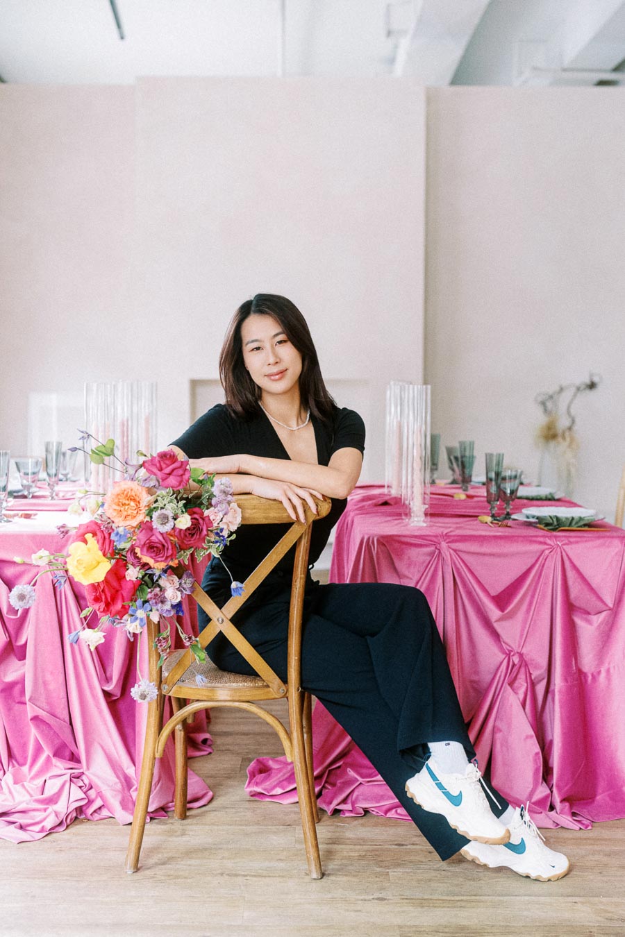 Elegant woman seated on a wooden chair against a pink-satin adorned table, with a vibrant floral arrangement, creating a chic, modern dining atmosphere.