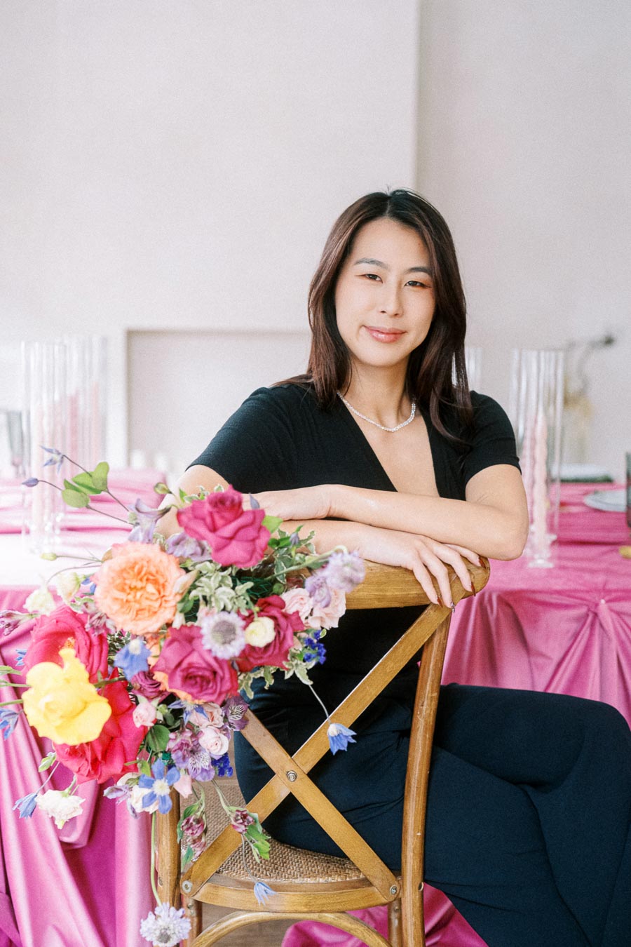 Smiling woman in a black outfit sitting on a wooden chair with a colorful bouquet of flowers, in a room with pink tablecloths and elegant decor.