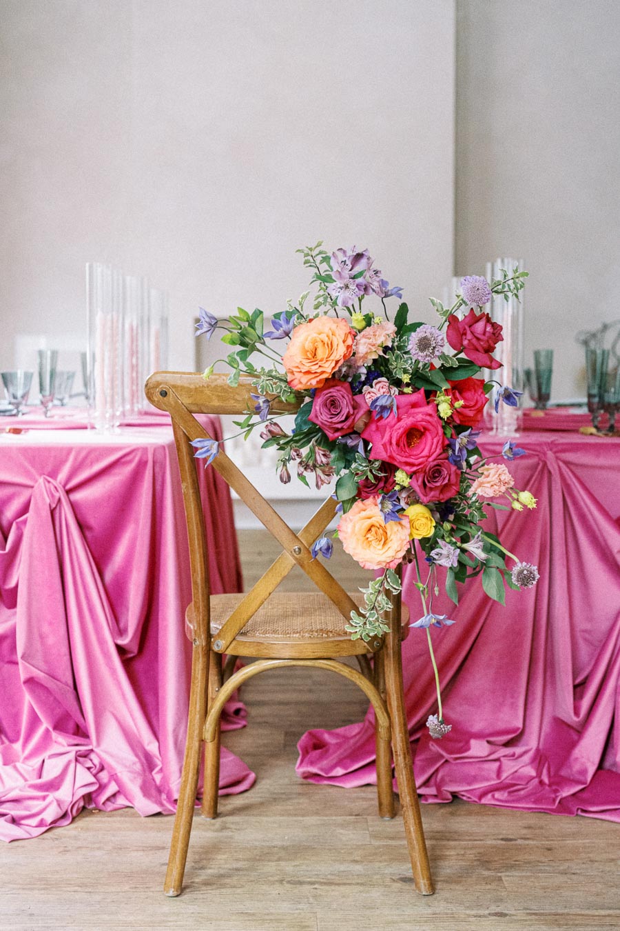 Wooden chair adorned with vibrant floral arrangement featuring pink, orange, and purple flowers, placed near a table draped with pink fabric, perfect for a stylish wedding or event setting.