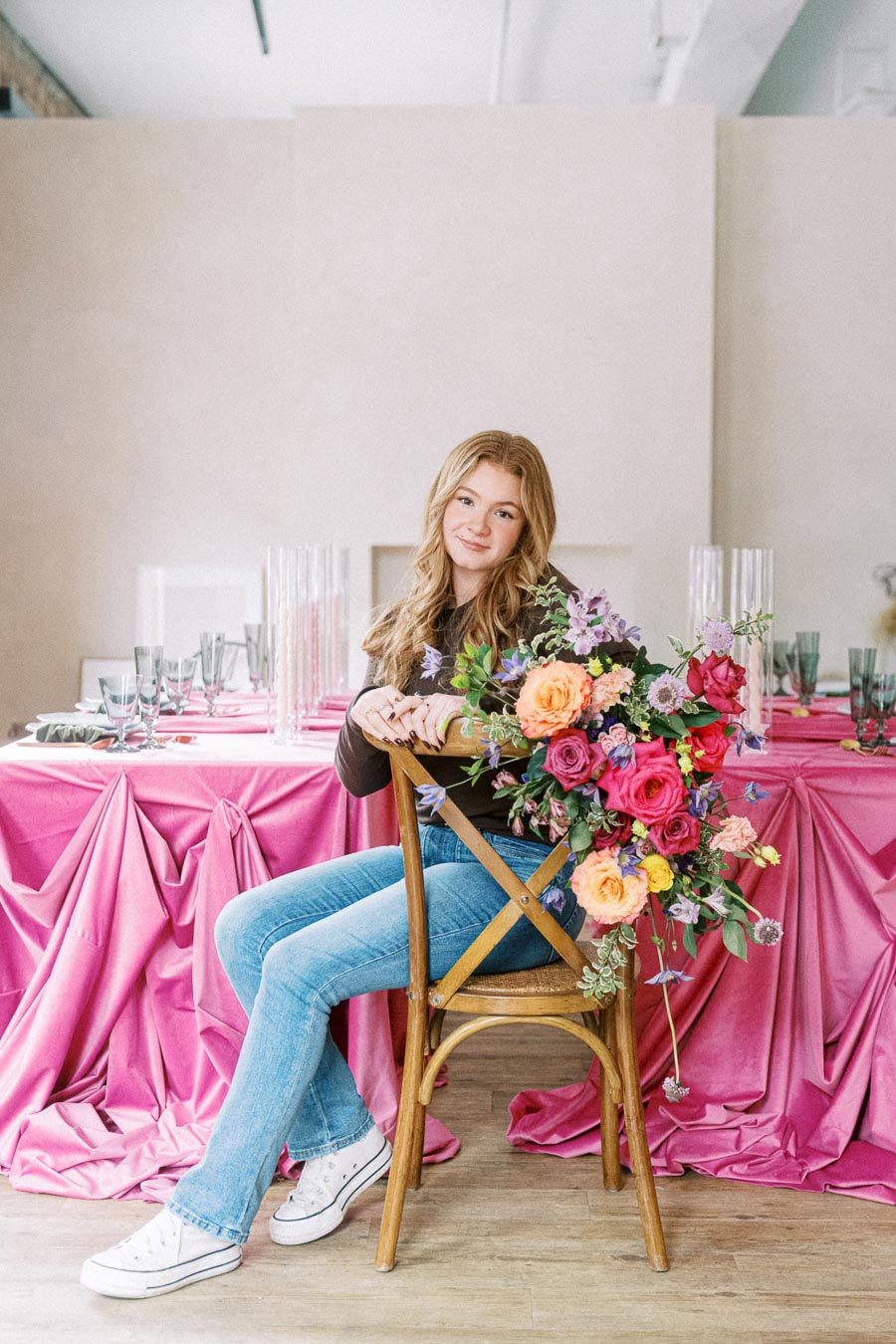 Young woman sitting on a wooden chair in front of a table with a vibrant pink tablecloth, adorned with an elegant floral arrangement in orange, pink, and purple hues. She is wearing casual jeans and white sneakers, with a bright indoor setting around her.