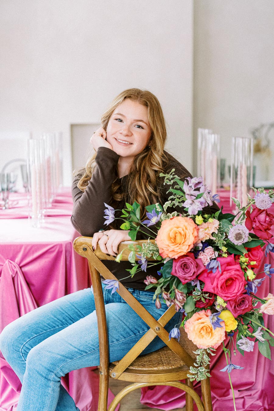A woman seated on a wooden chair next to a vibrant floral arrangement, featuring pink, orange, and purple flowers, in front of a table with a pink tablecloth, creating an elegant and cheerful ambiance.