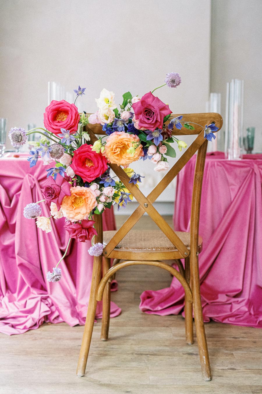 Wooden chair adorned with a colorful floral arrangement featuring pink, purple, and orange flowers, set against tables covered with pink fabric, ideal for a vibrant wedding or event decoration.