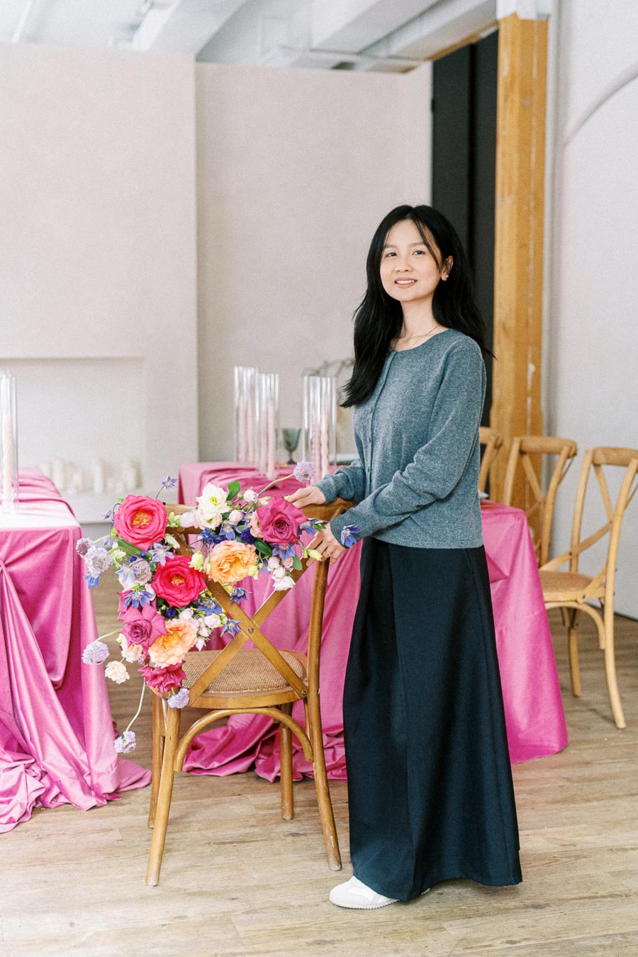 Woman standing next to a wooden chair adorned with vibrant floral arrangement in a room with pink-draped tables and elegant decor.