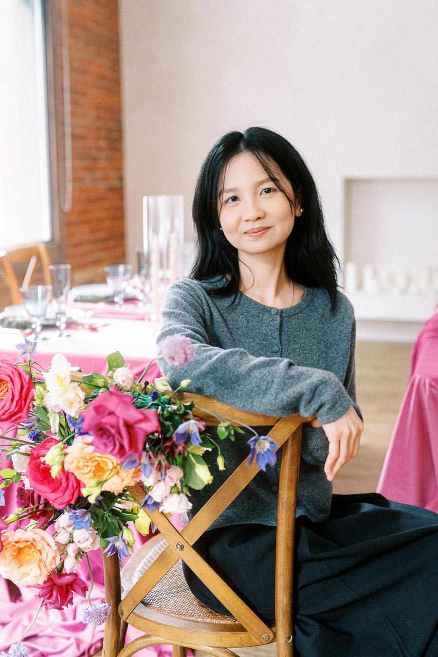 Young woman sitting on a wooden chair adorned with vibrant flowers, in a tastefully decorated dining room with pink tablecloths and elegant table settings.