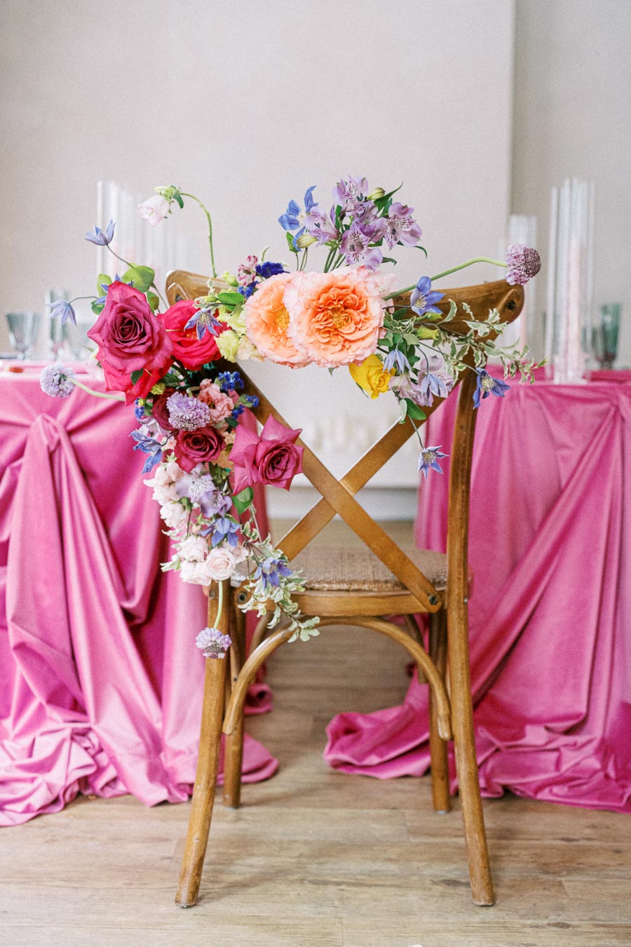 Rustic wooden chair adorned with vibrant floral arrangement featuring pink, purple, and orange flowers, set against a stylish pink draped table in a wedding or event setting.