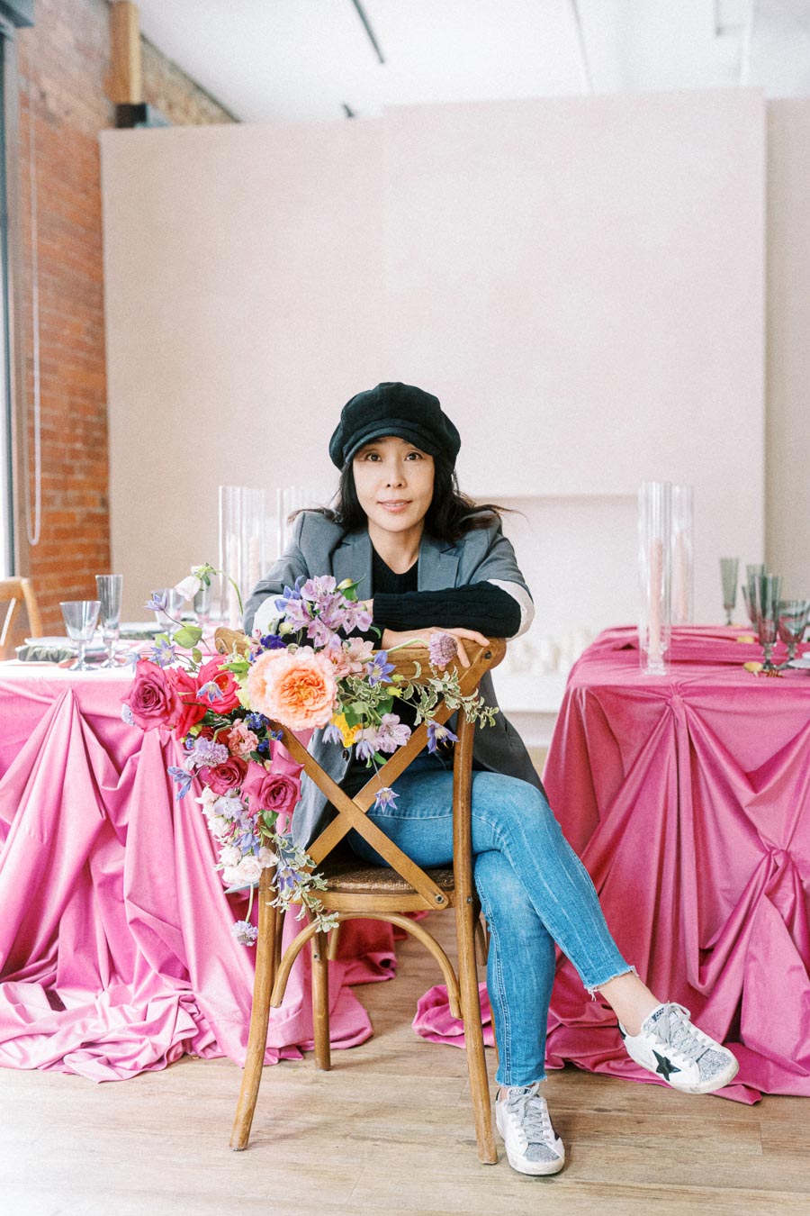 A woman sitting on a chair adorned with vibrant flowers, surrounded by elegantly set tables with pink tablecloths in a stylish interior space.