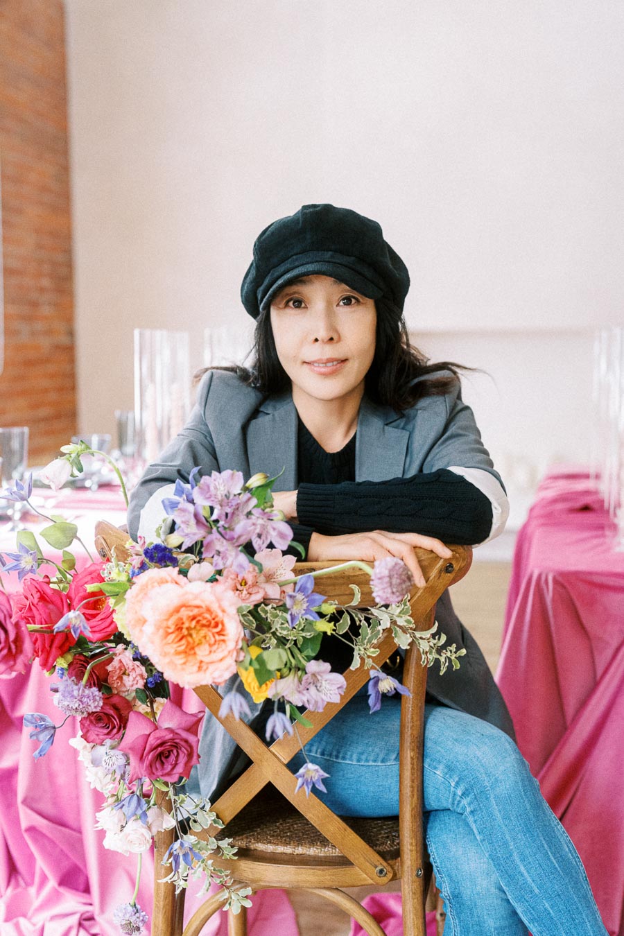Woman in a black hat sitting on a wooden chair adorned with vibrant flowers, set against a backdrop of pink table covers in a stylish interior space.