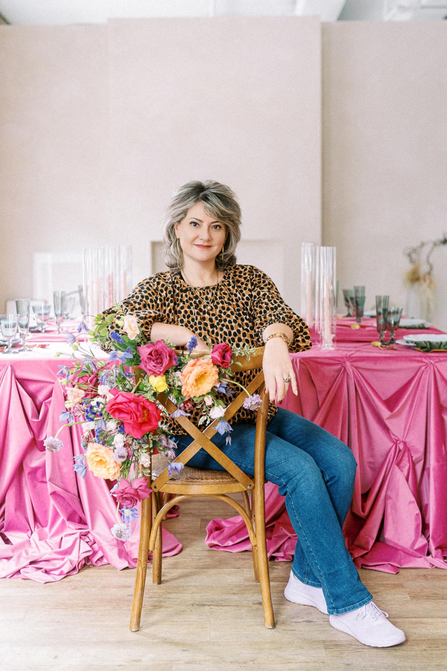 Woman seated on a wooden chair adorned with colorful flowers, in front of a pink draped table setting with glassware and cutlery, wearing a leopard print blouse and jeans.