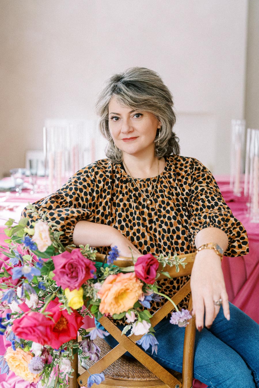 A woman with short hair wearing a leopard print top sits on a wooden chair adorned with colorful flowers, set against a background with a pink tablecloth.