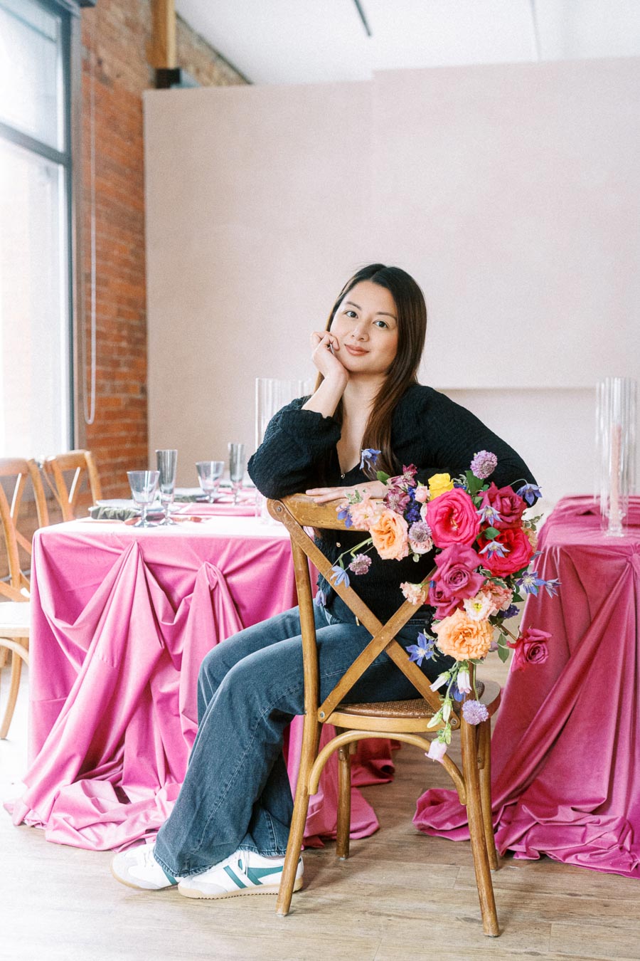 Woman seated at a table with vibrant pink floral arrangement and draped pink tablecloth, in a cozy, well-lit room with brick walls.