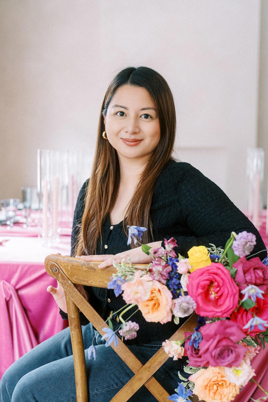 A woman sitting on a wooden chair with colorful flowers in the foreground and a table set with pink tablecloths in the background, creating an elegant event setting.