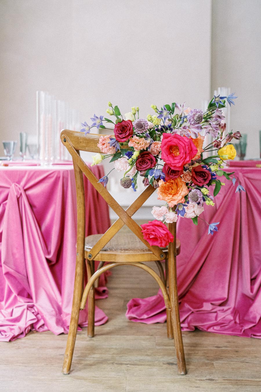 Elegant wedding reception setup with a wooden chair adorned with vibrant floral arrangement, featuring pink, orange, and red flowers, next to a table draped in luxurious pink fabric.