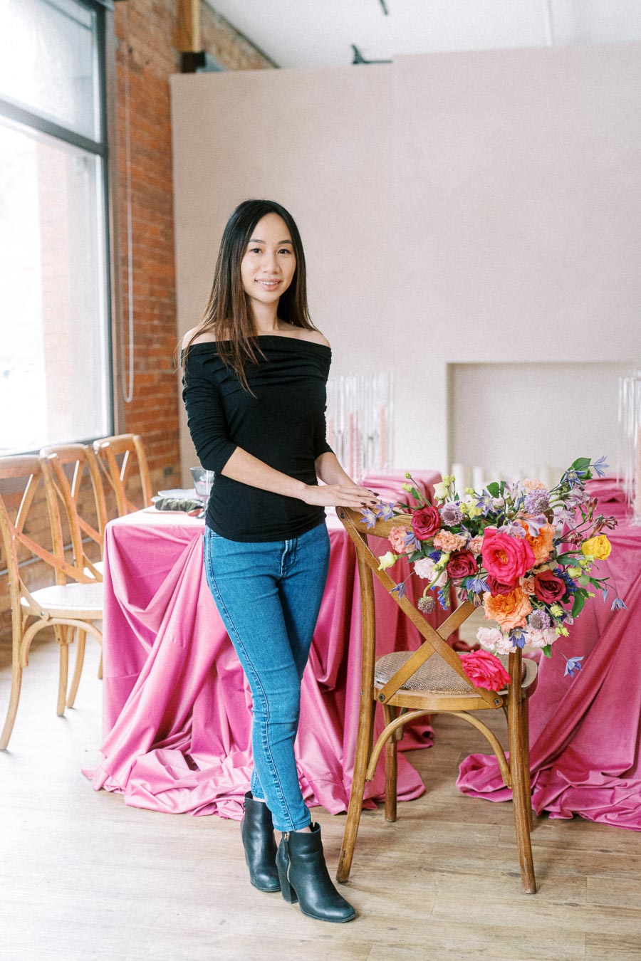 A woman in a black top and jeans stands in a stylishly decorated room with pink draped fabric and a vibrant floral arrangement on a wooden chair.