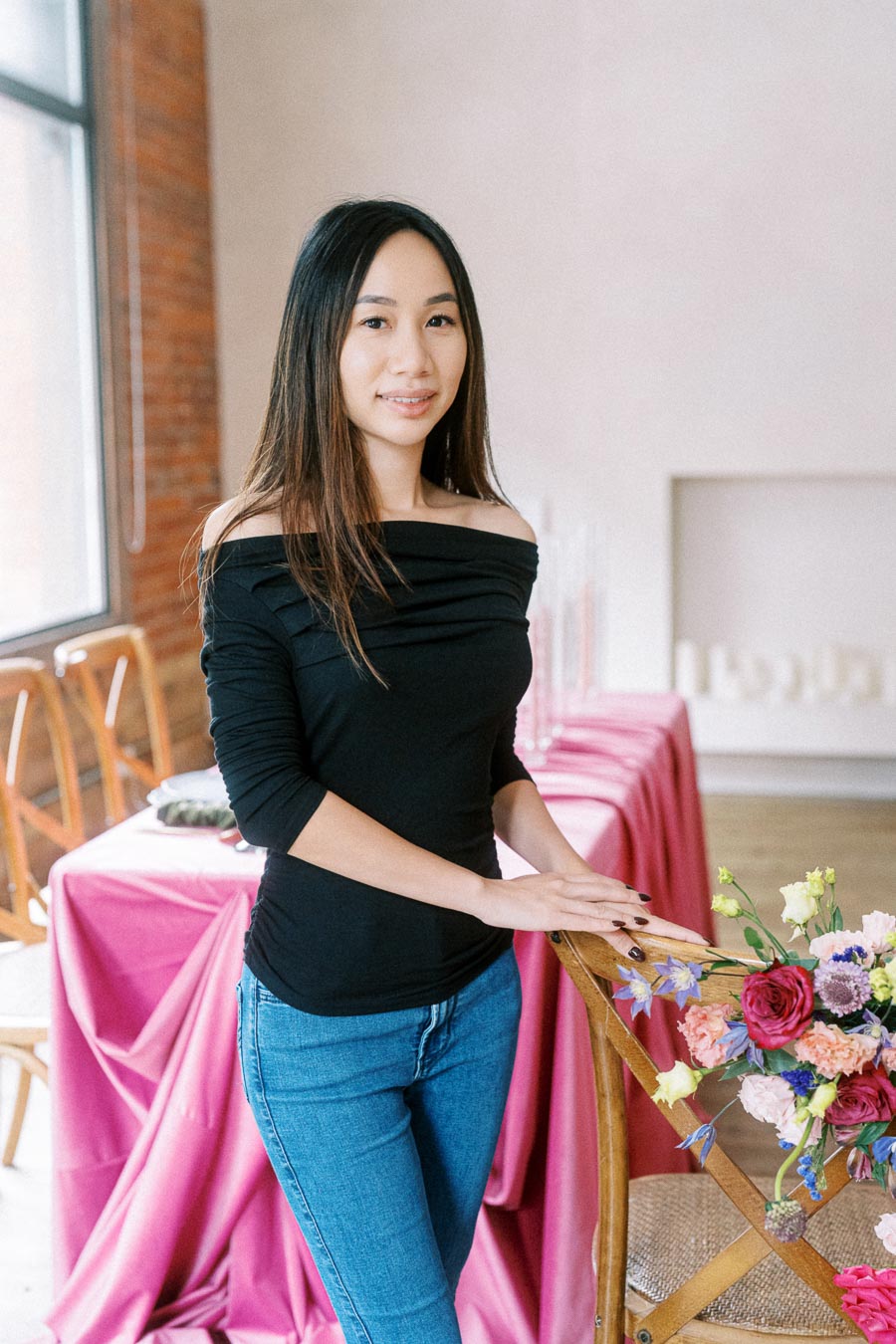 Smiling woman in a black off-shoulder top and jeans standing beside a wooden chair with colorful floral arrangement in a bright room with pink tablecloths.