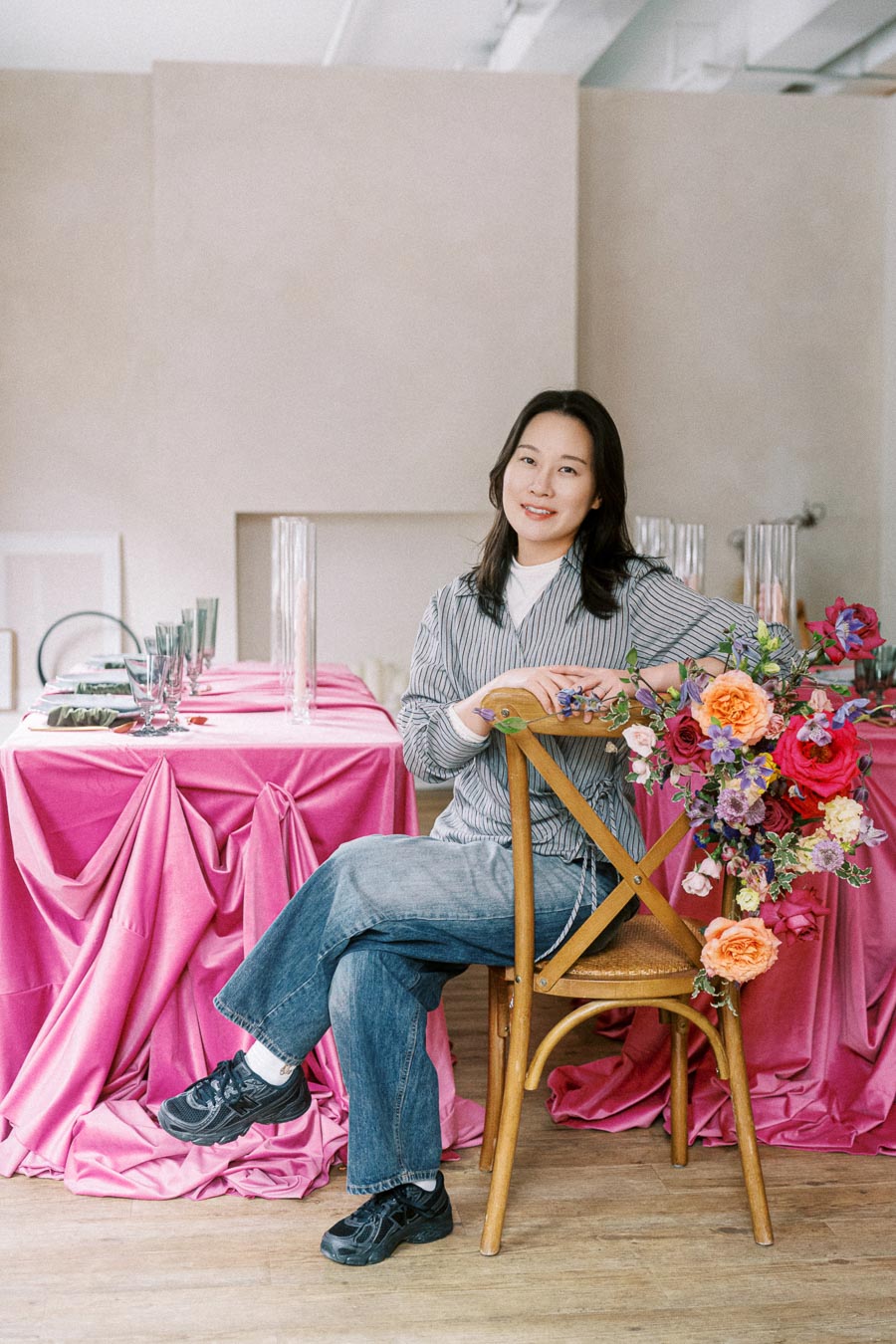 A woman seated on a wooden chair beside a table draped in vibrant pink fabric, adorned with a colorful floral arrangement. She is wearing casual attire, including a striped blouse and denim jeans, and the setting has a serene and stylish ambiance.