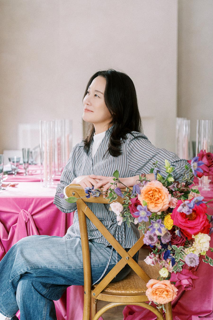 A woman in a striped shirt sits on a wooden chair decorated with colorful flowers, surrounded by a table elegantly set with pink tablecloths and glassware.