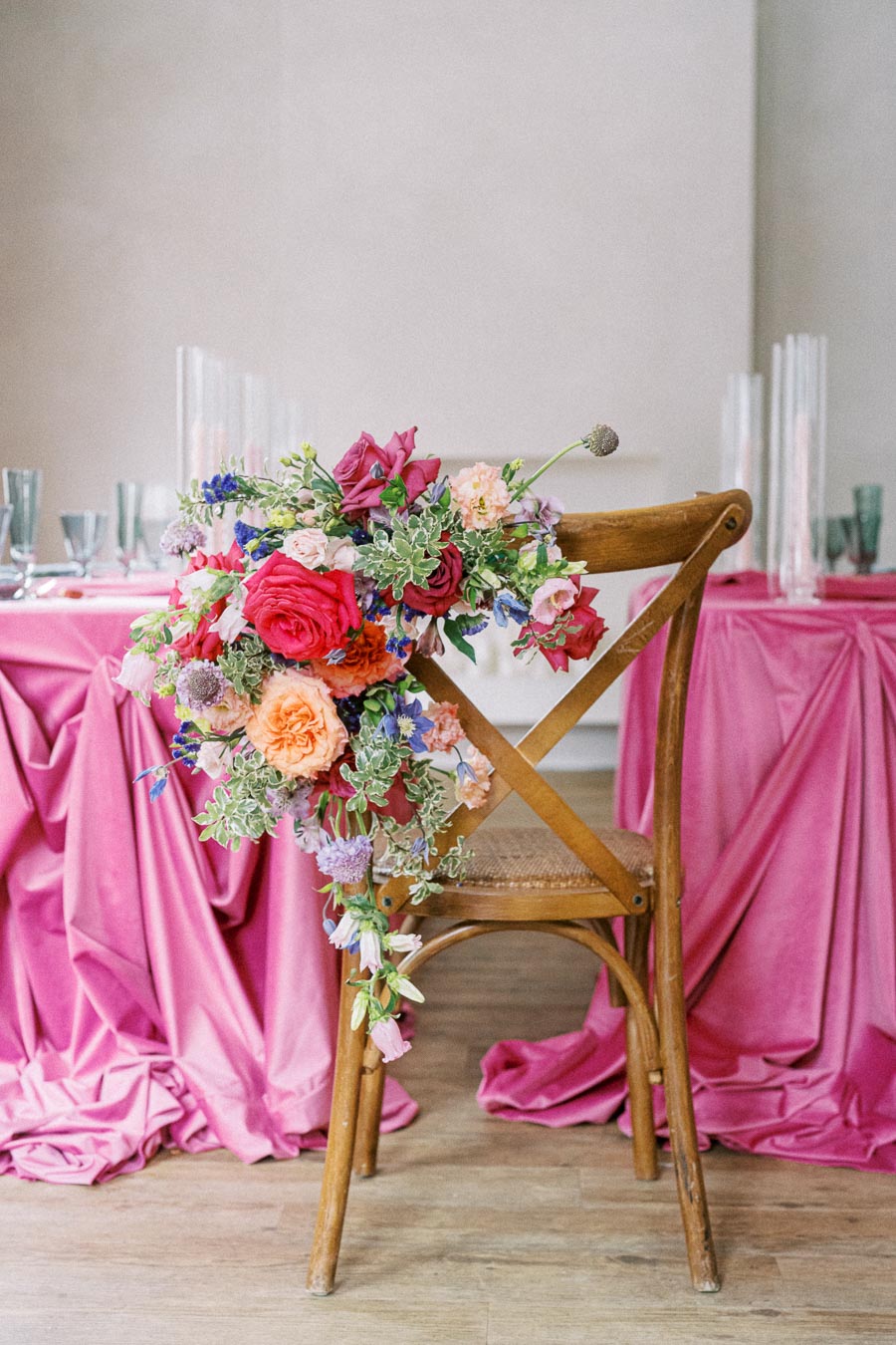 Elegant wooden chair adorned with vibrant floral arrangement, featuring red, pink, orange, and purple flowers, set beside a table draped in luxurious pink fabric for a romantic celebration setting.