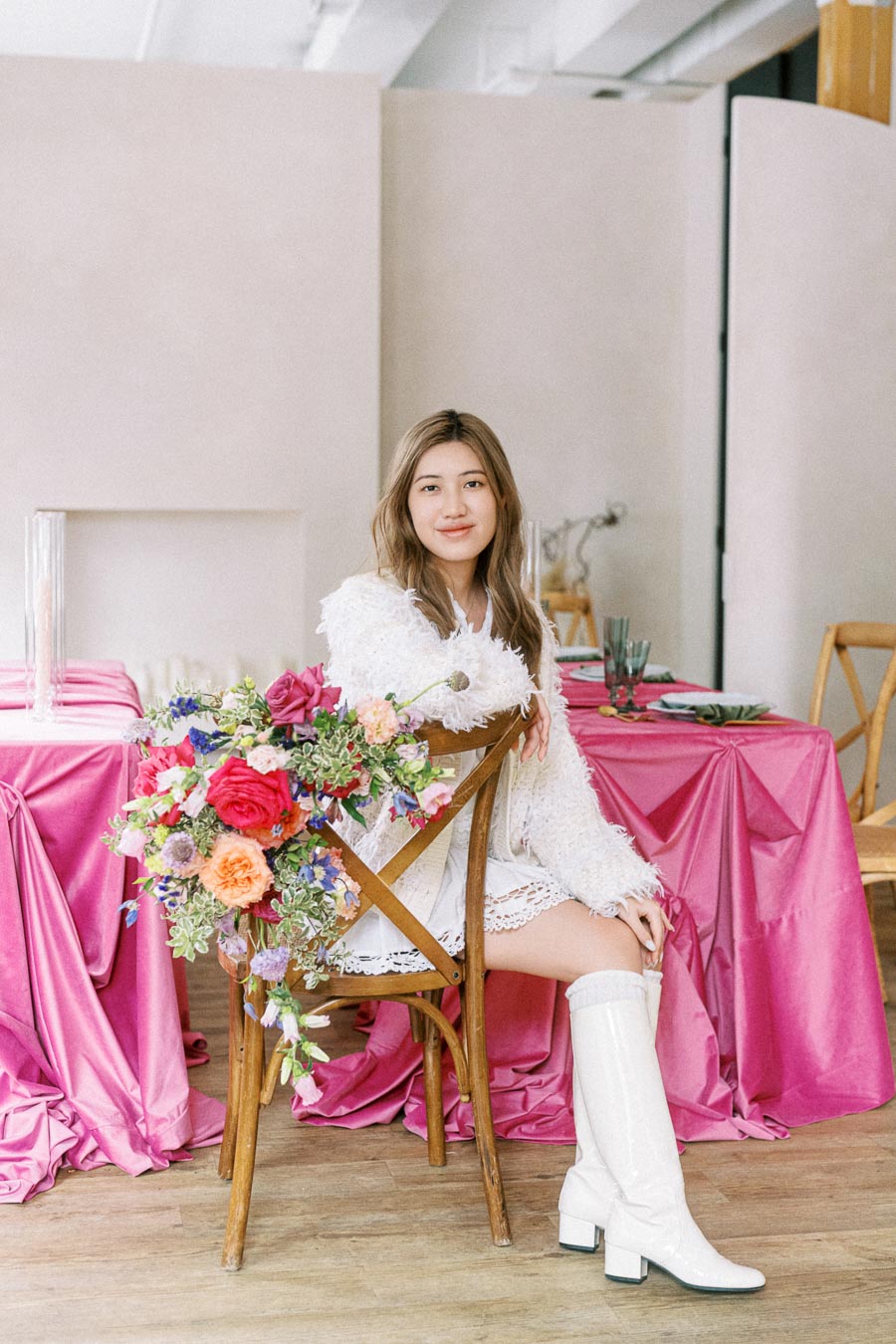 A woman in a white dress and boots sits on a wooden chair adorned with colorful flowers, next to a table with pink satin tablecloths in a stylish interior setting.