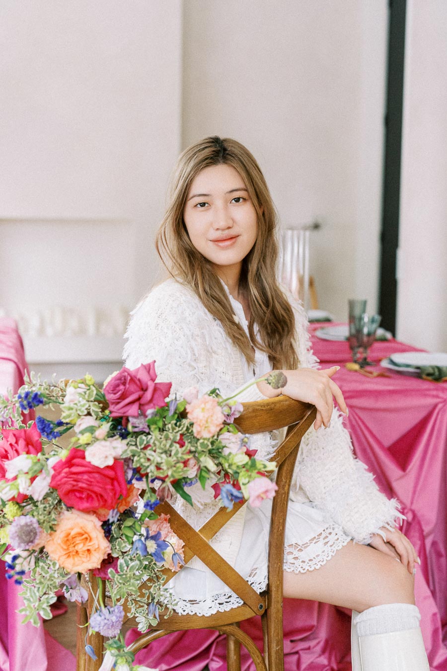 A woman in a white outfit sitting on a wooden chair with a vibrant bouquet of flowers attached to it, set in a room with pink decor and elegantly arranged dining table settings.