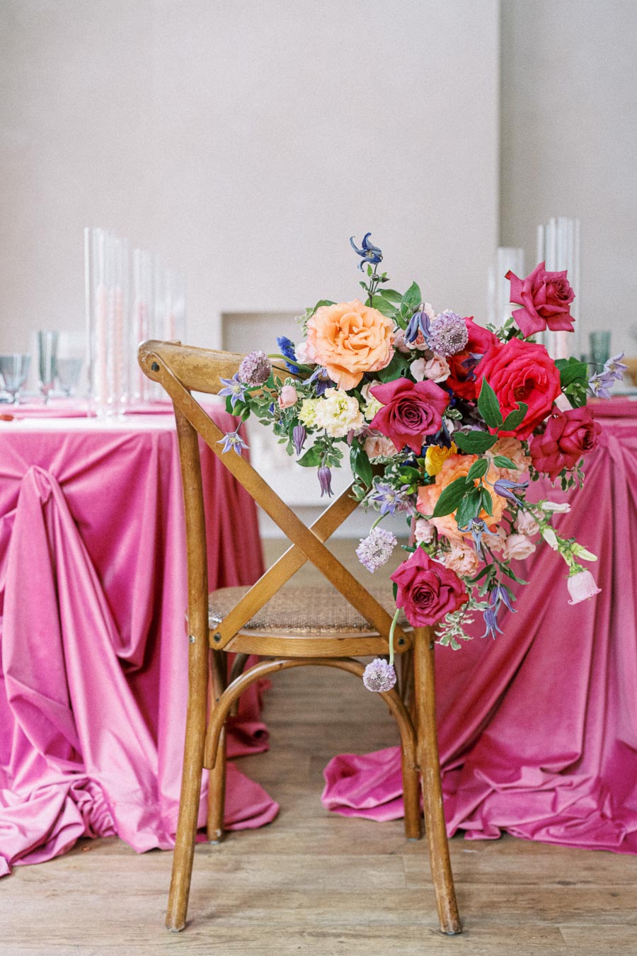A wooden chair adorned with a vibrant floral arrangement featuring red, pink, and orange roses, set against a pink draped tablecloth, creating an elegant and romantic event setting.