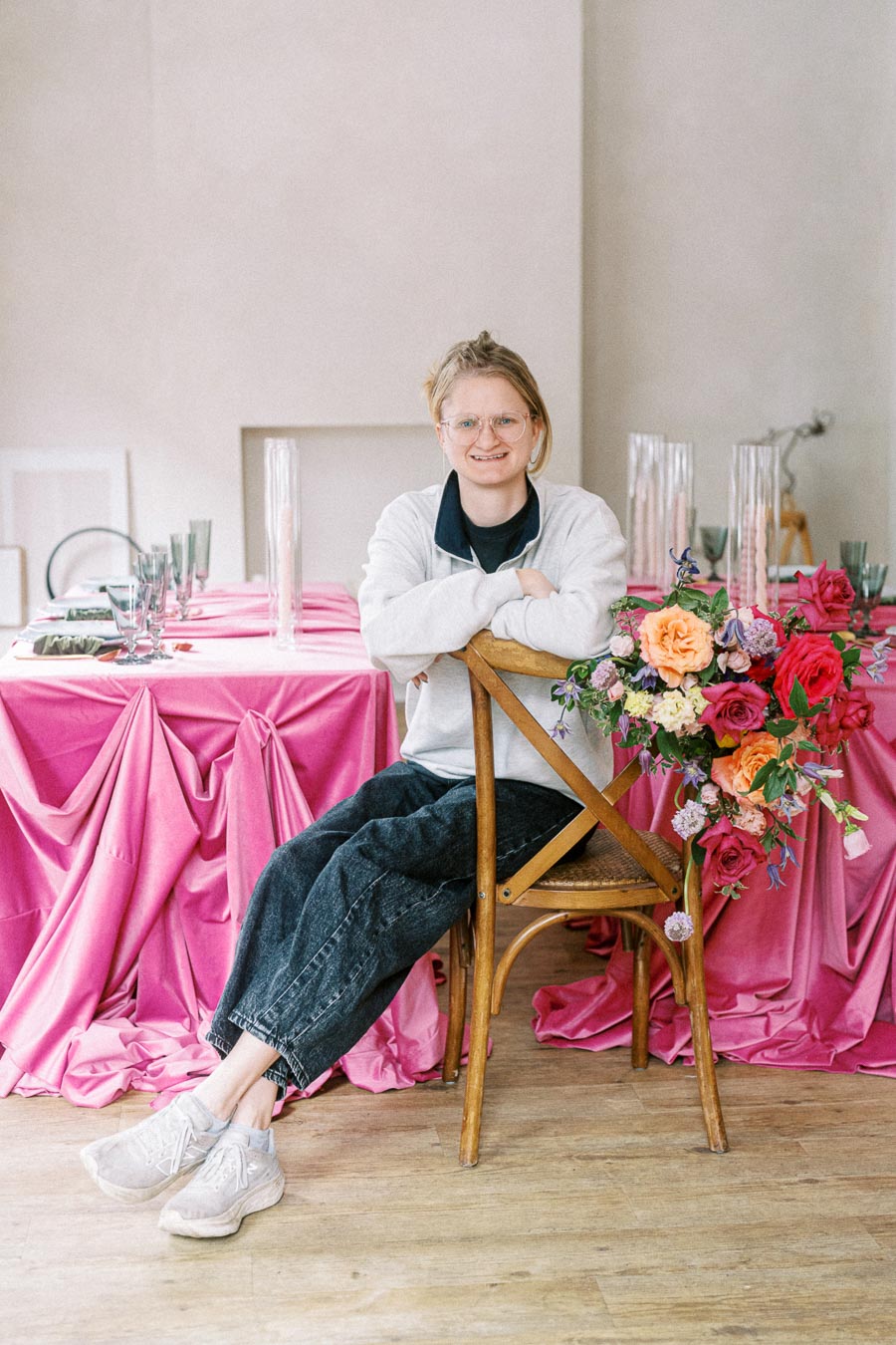 Person sitting on a wooden chair at a beautifully decorated table with pink fabric and a vibrant floral arrangement, showcasing a stylish event setup.