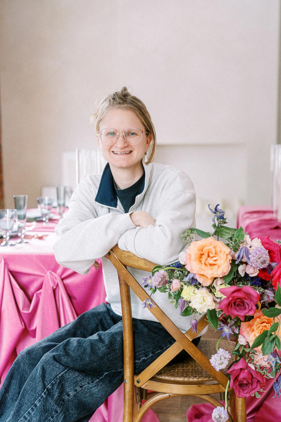 A person sitting on a wooden chair with a colorful floral arrangement, smiling and wearing glasses. A table with a bright pink tablecloth and glassware is in the background, suggesting a celebratory or event setting.