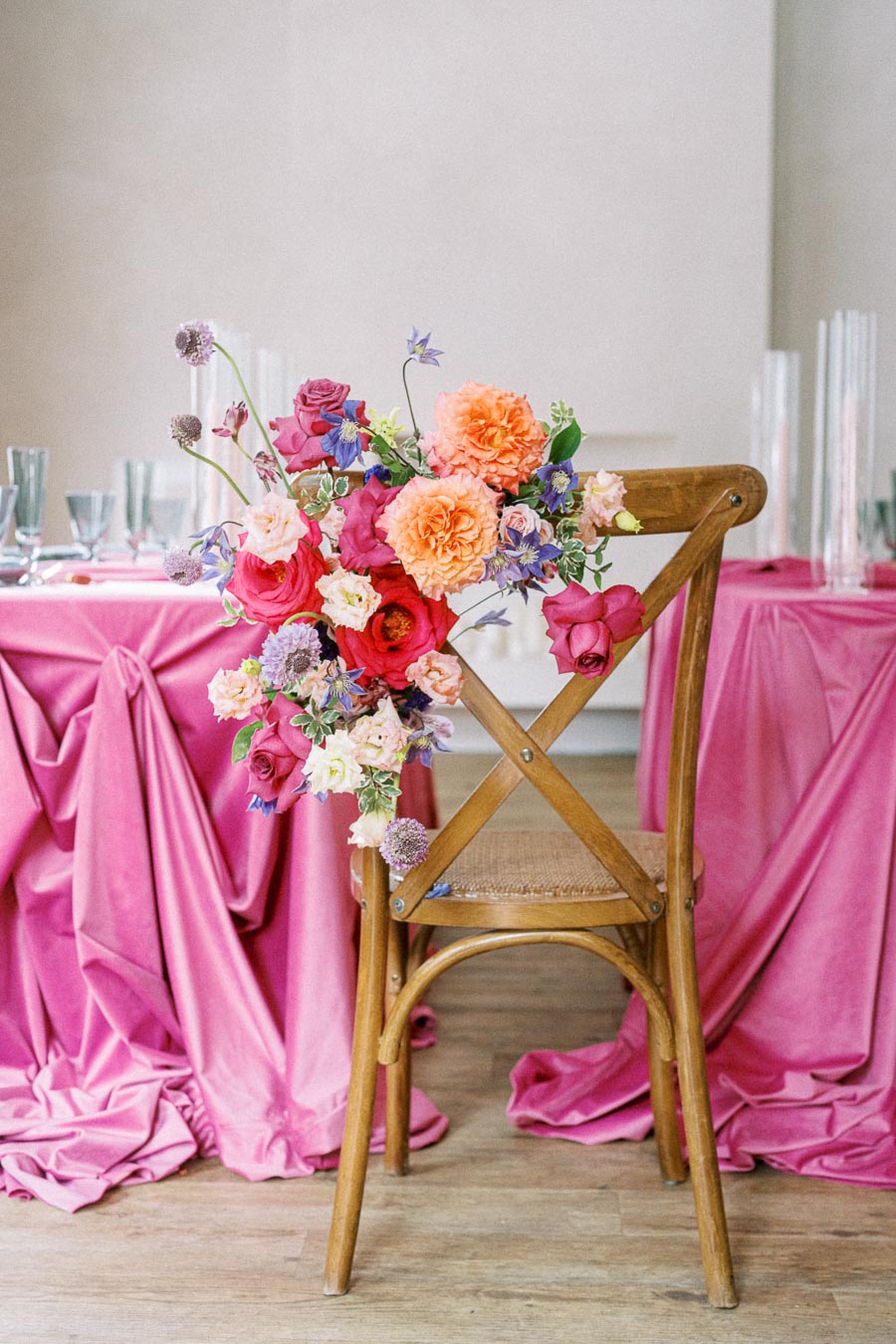 Wooden chair with vibrant floral arrangement, including pink, orange, and purple flowers, set against a backdrop of pink draped tablecloths and glassware in an elegant event setting.