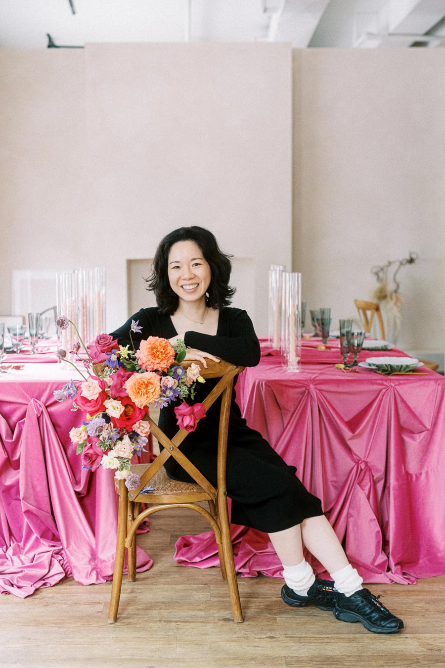 A smiling woman sits on a wooden chair adorned with colorful flowers. The background features a table draped with vibrant pink fabric and set with elegant glassware. The setting exudes a cheerful and welcoming atmosphere, ideal for celebrating special occasions.