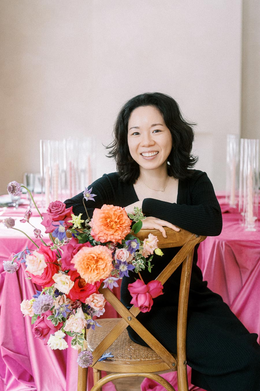 A person smiling while sitting on a wooden chair with colorful flower arrangement, against a backdrop of a pink tablecloth setting.