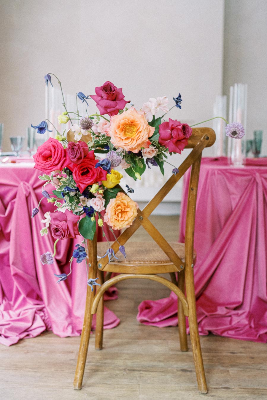 Elegant wooden chair adorned with a vibrant floral arrangement, featuring pink, orange, and yellow roses, set against a backdrop of rich pink tablecloths, creating a romantic and colorful wedding or event setting.