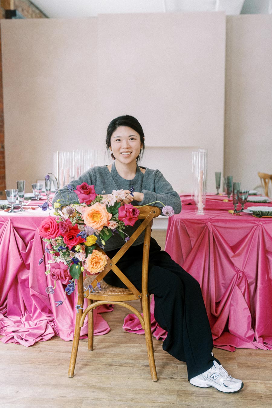 Young woman sitting on a chair with vibrant floral arrangement, smiling at a table elegantly draped in pink fabric, decorated with glassware, in a cozy, well-lit setting.