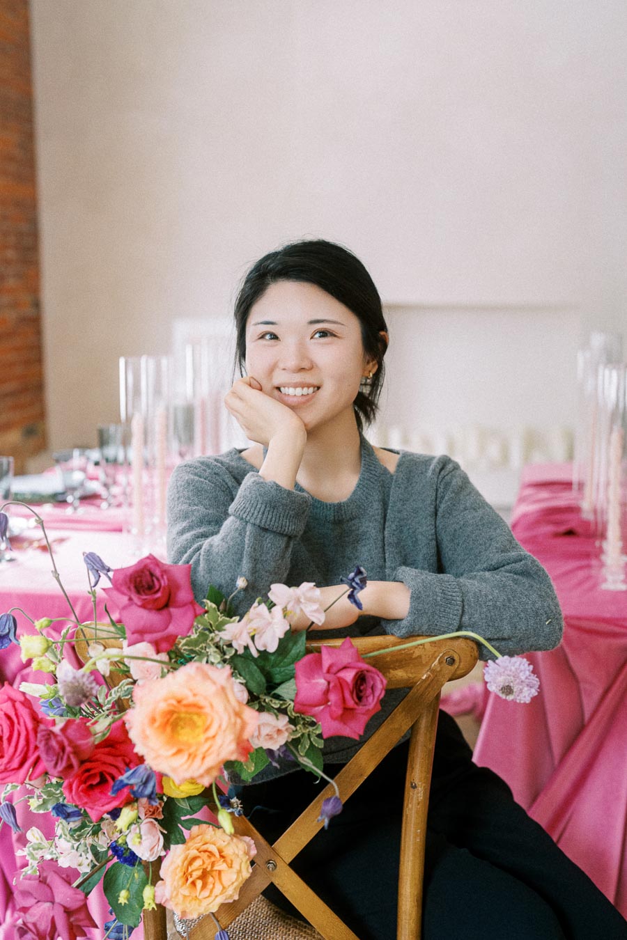A woman smiling while sitting at a table decorated with vibrant pink and orange flowers, dressed in a cozy gray sweater, with a warm and inviting atmosphere in the background.