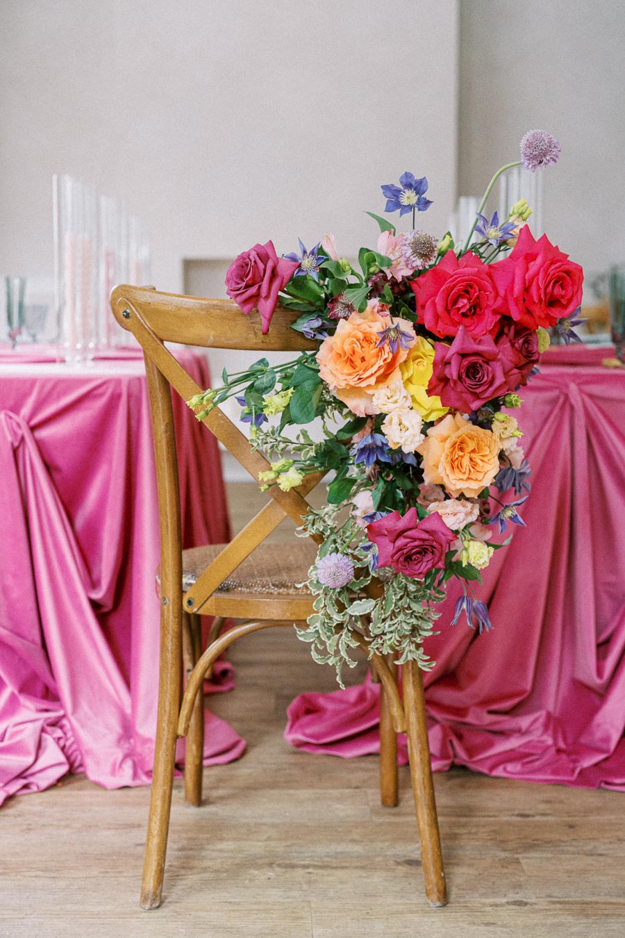 Wooden chair adorned with a vibrant floral arrangement featuring red, pink, orange, and yellow roses, placed beside a dining table draped in pink fabric, embodying elegant wedding decor.