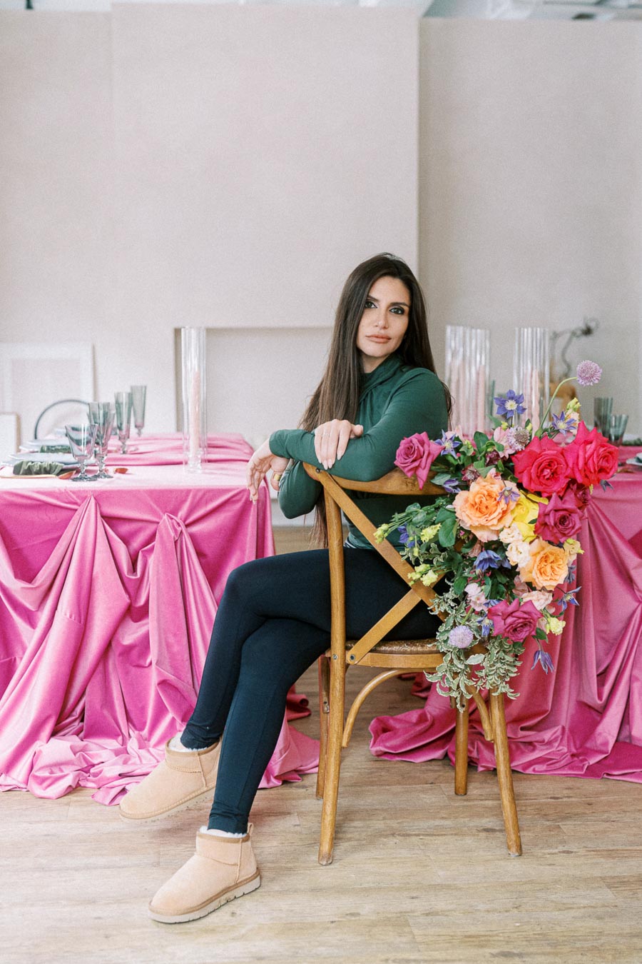 A woman in a green long-sleeve top sits on a wooden chair adorned with colorful flowers, set beside a table covered in pink fabric. The setting includes neatly arranged glassware, creating a stylish and vibrant atmosphere.
