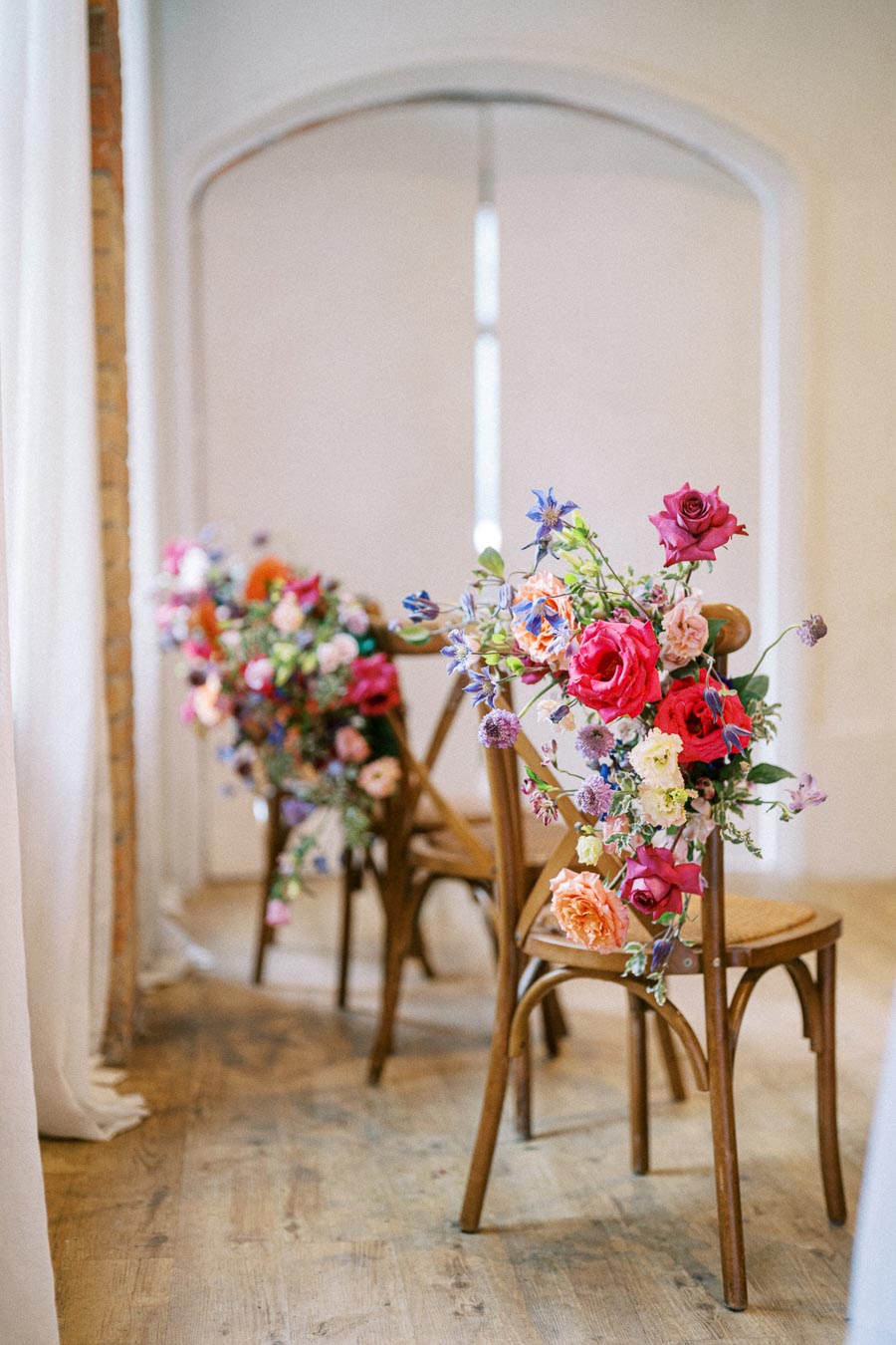 Elegant wooden chairs adorned with vibrant floral arrangements, featuring pink, red, and orange flowers, in a bright, airy room with soft natural light.