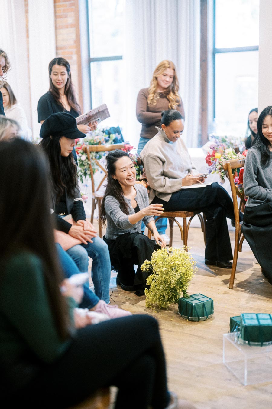 Group of people participating in a floral arrangement workshop, surrounded by colorful flowers and greenery, in a well-lit room with large windows.