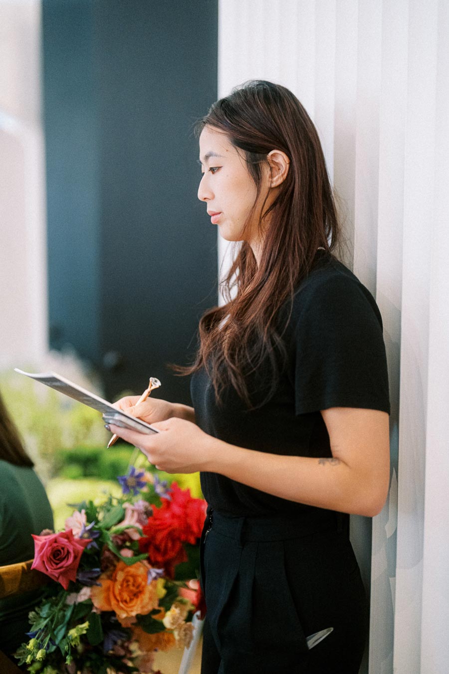 Young woman in a black shirt taking notes while standing against a white wall, with a colorful bouquet of flowers in the background.