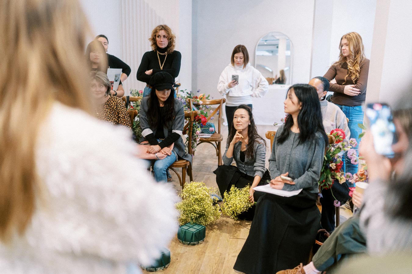 A diverse group of people gathered in a bright room, engaged in a workshop or meeting. Some are seated with notepads, others are standing, and plants decorate the space, creating a creative and collaborative atmosphere.