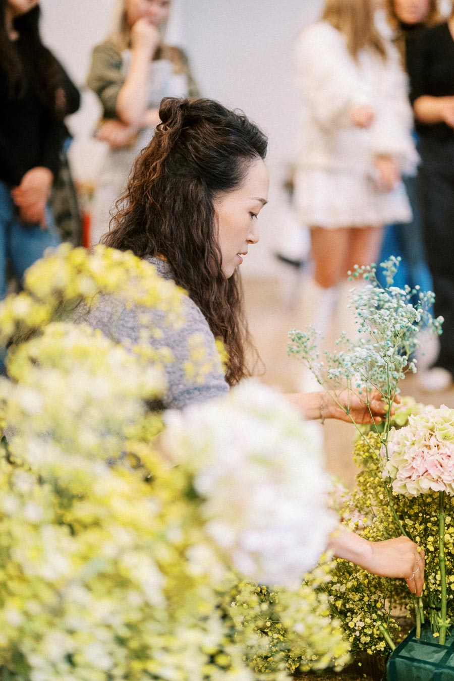 A florist arranging a bouquet of colorful flowers at a workshop, surrounded by people observing in the background.