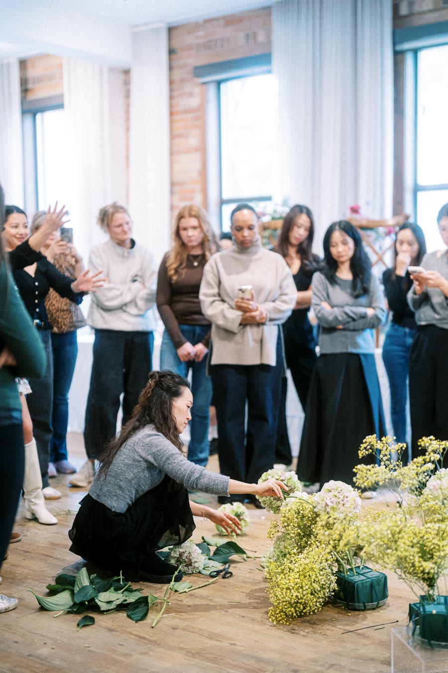 A group of people attentively watching a female florist in a workshop as she arranges colorful flowers on a wooden floor, surrounded by vibrant green leaves and stems in a well-lit room with large windows.