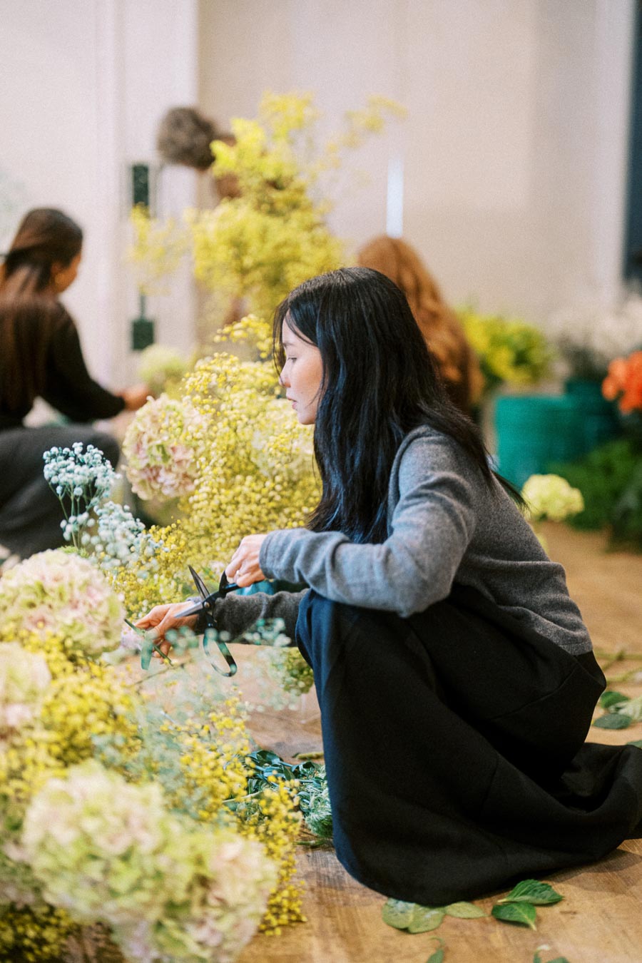 A woman arranging flowers with scissors in a floral workshop, surrounded by various colorful blooms and greenery.