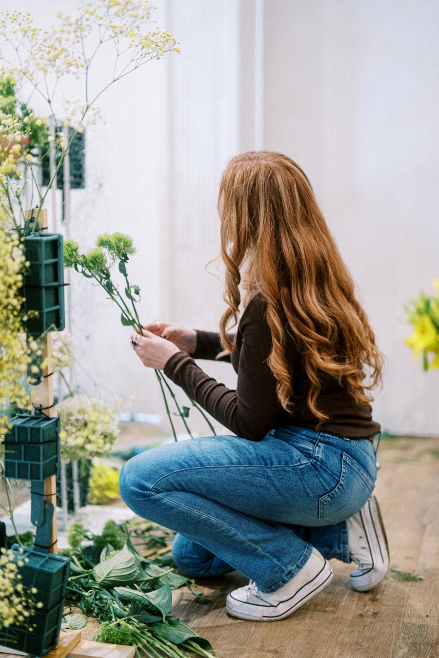 A woman with long red hair arranges green stems and flowers while kneeling on a wooden floor, showcasing a floral design process in a creative workspace.